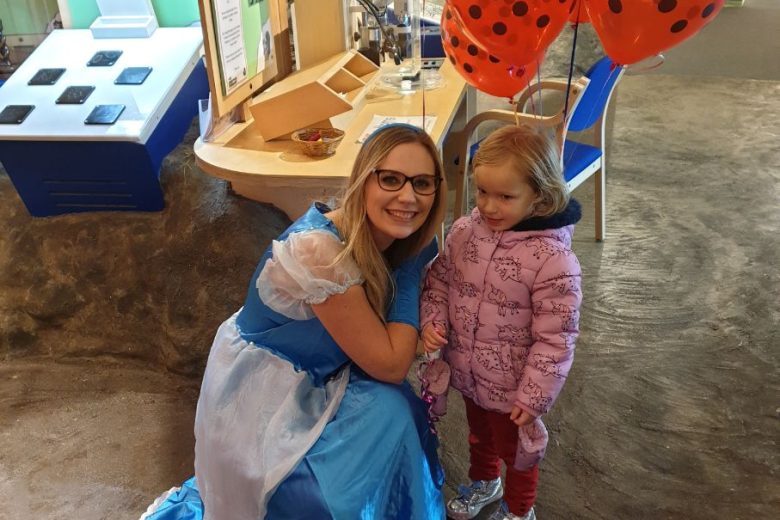 Participation worker dressed up as Cinderella. She is knelt down next to a young girl holding a ladybird balloon.