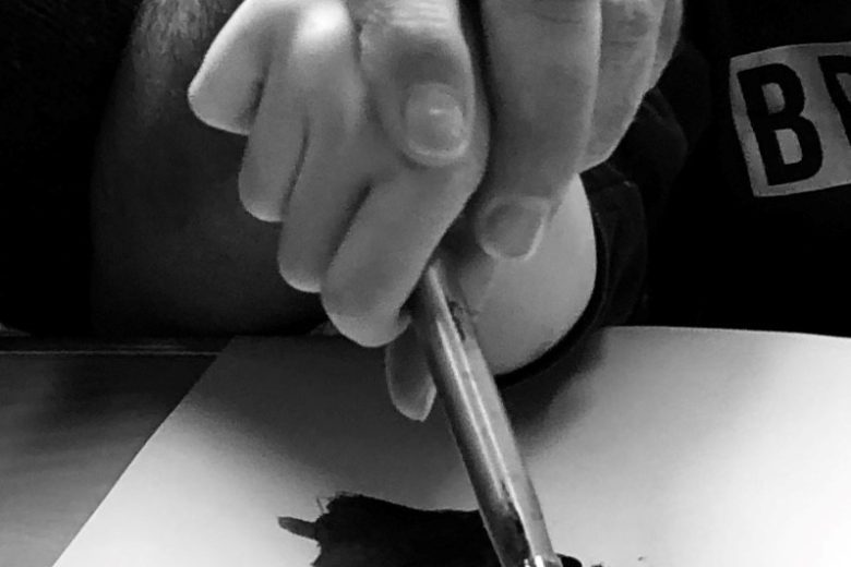 Black and white photo of one hand on top of another holding a paintbrush. Painting is a partially visible black shape.