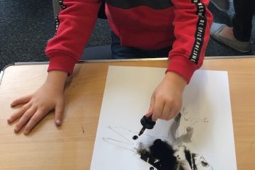 Child's hands, one holding a paintbrush to a black watercolour painting of a bird