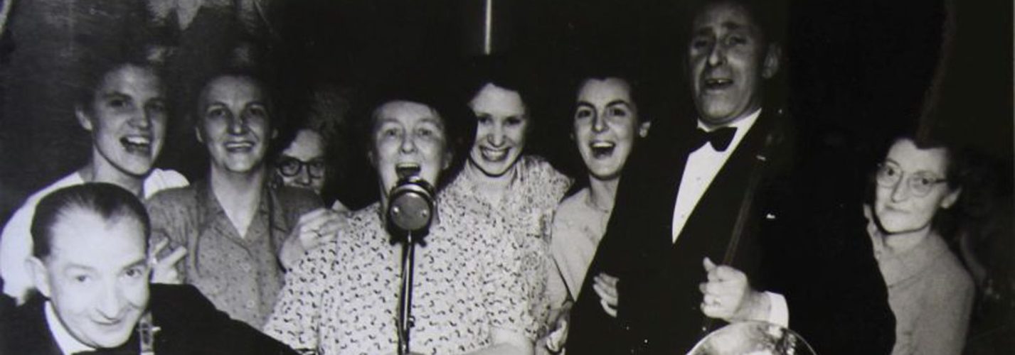 Old black and white photo of adults dressed formally and smiling at the camera. One woman is singing into an old fashioned microphone.