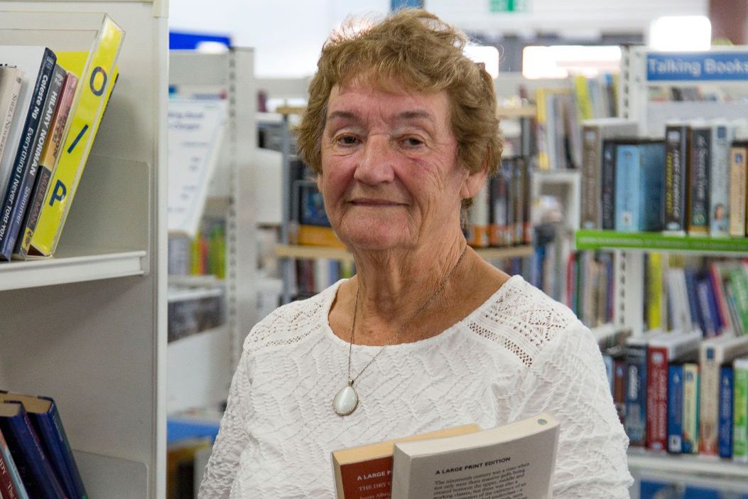 Volunteer stood in a library holding some books and looking at the camera.