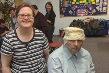 Woman smiling next to man sat down with a bandage around his head