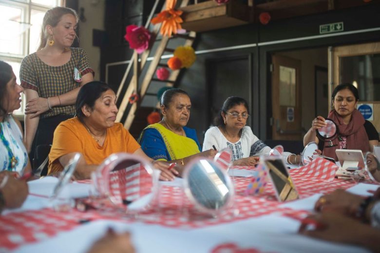 Workshop leader stood at a table with a group of ladies sat down. They are using mirrors to help them stitch their self portraits