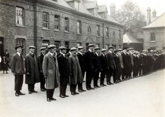 Black and white photo of a group of men wearing long coats and flat caps lined up in two rows