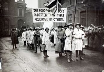 Black and white photo of women marching. they are holding a sign that says the food you produce is better than that produced by others