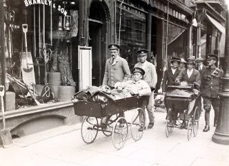 Black and white photo of soldiers walking down the street. one is being pushed in a pram.