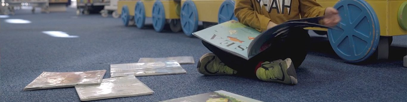 Image of a child sat on the floor reading. Only the lower half of the child is visible. Three other books are on the floor.