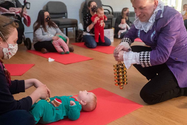 Woman in a mask and a performer holding a sleigh bells instrument smiling at a baby lying down on a mat.