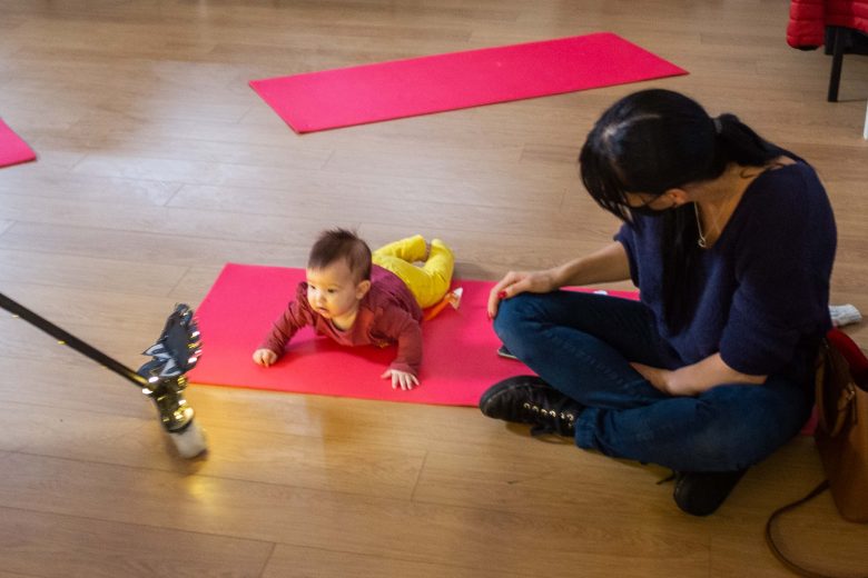 Woman wearing a mask looking down at a baby lying on a mat and looking at the broomstick toy.