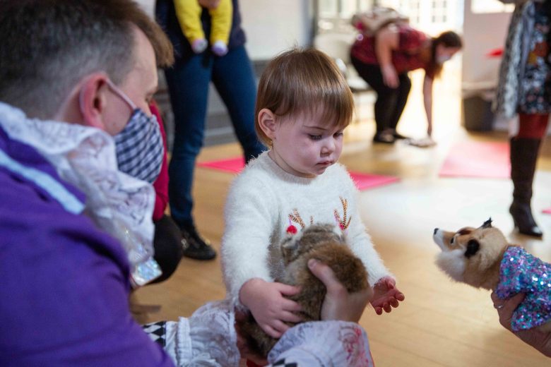 Performer and woman both wearing masks and holding out stuffed toys to a small child