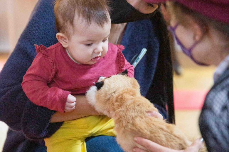 Women wearing masks, one with a baby on her lap and the other holding out a stuffed animal to the baby