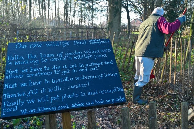 Information board that reads: our new wildlife pond started February 2020. hello, the team of garden volunteers is working to create a wildlife pond. first we have to dig it out in a shape that allows creatures to get in and out. next we have to install a waterproof lining. then we fill it with water! finally we will put plants in and around the pond. we are following good advice in a book on ponds.