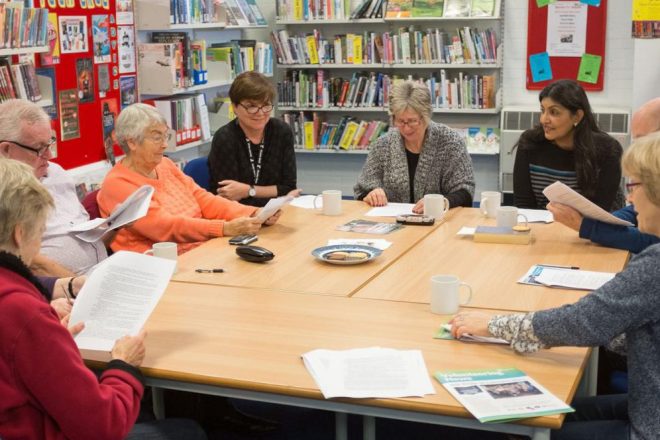 Group sat around a library table with coffee and biscuits. They are reading printed pages with text on them.