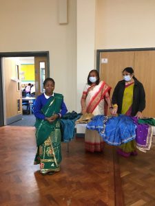 School child wearing a sari over her uniform. Two anand mangal ladies are stood behind her at a table with different coloured fabrics on it.