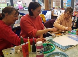 A group of women sat around a table and painting on a small canvas.