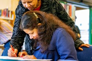 A woman sat at a table with her hands on a piece of paper. Another woman is leaning over her and holding a pen to the page.