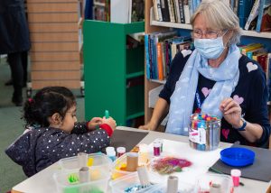 Woman wearing a mask sat at a crafts table with a young girl who is drawing