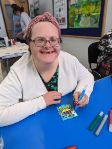 A woman with Down's syndrome smiling at the camera and colouring in a illustration