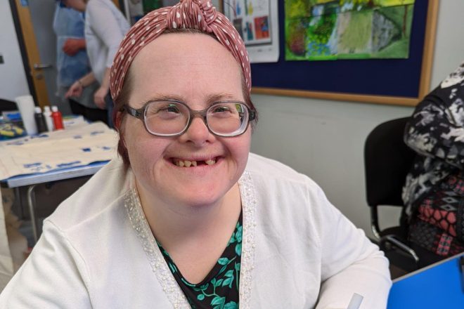 A woman with Down's syndrome smiling at the camera and colouring in a illustration