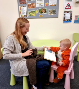 Woman and baby sat in little chairs. Baby is touching the page of a book.