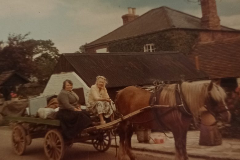 Old coloured photograph of two women sat in a horse-drawn wooden cart